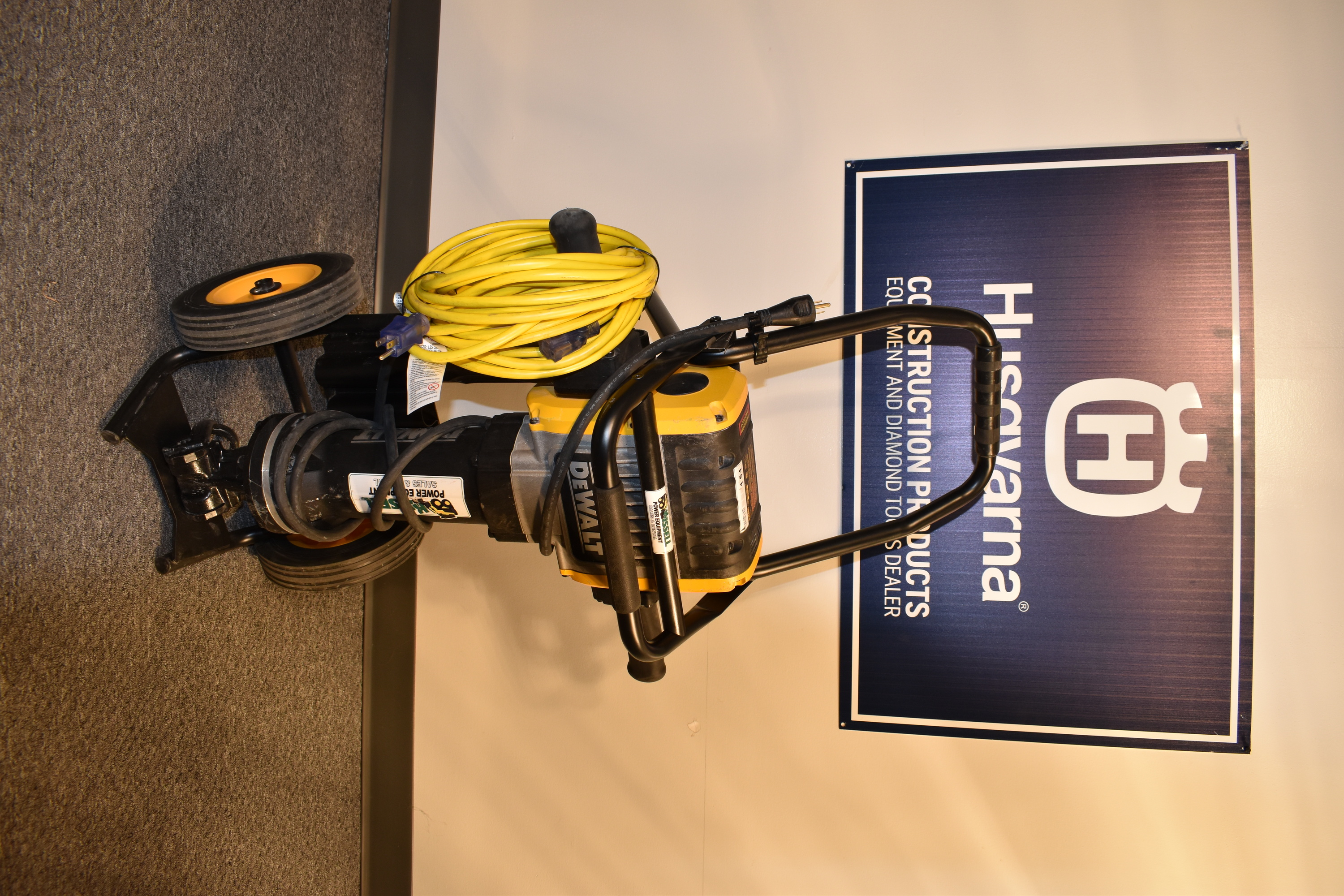 A dewalt electric jackhammer is propped upright on its stand. It has a long hammer, a large engine, and a yellow power cord wrapped around a handle. 