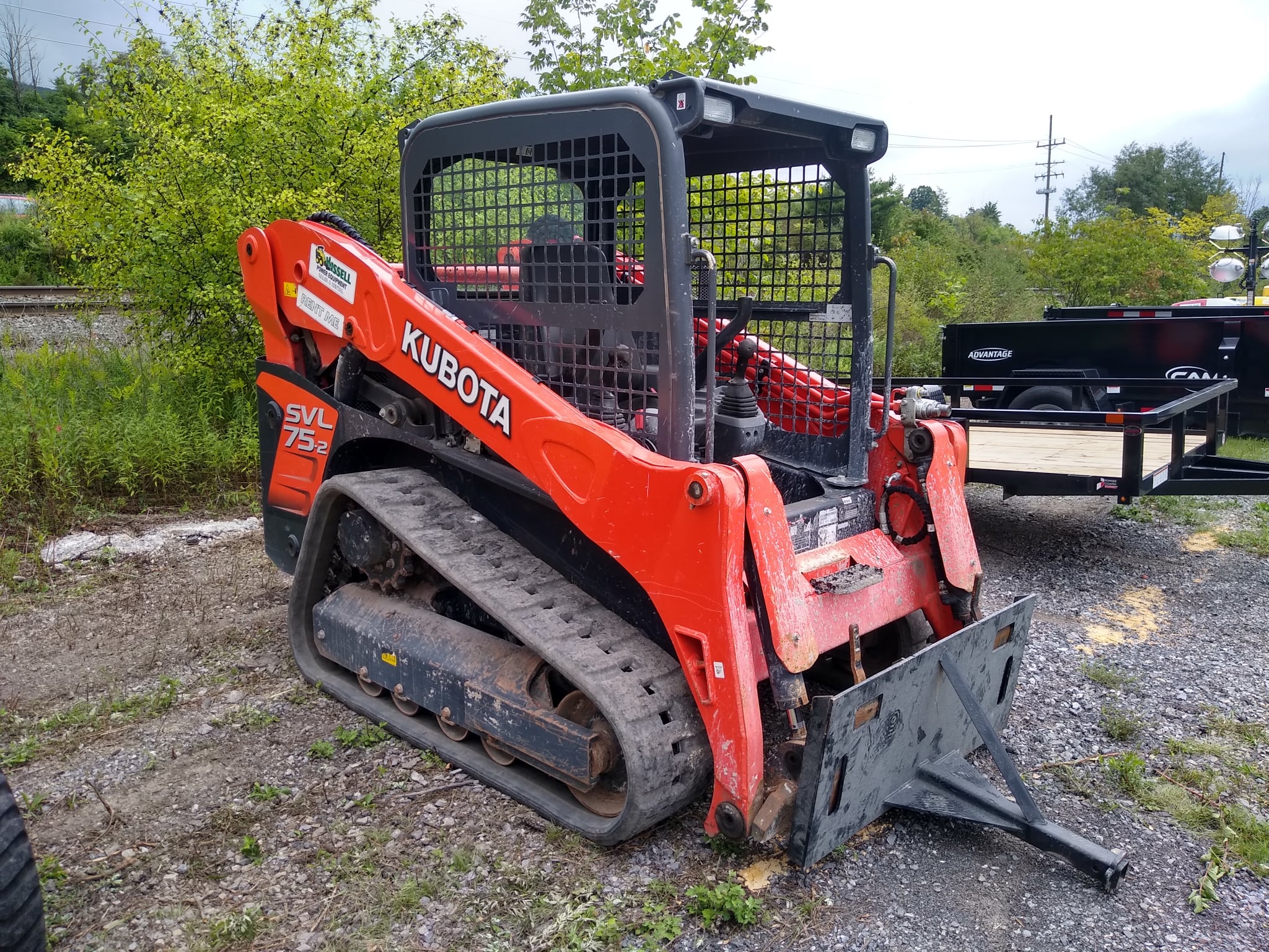 A Kubota skid steer with tracks is sitting in front of a row of bushes. The orange machine has an opening for attachments on the front. 