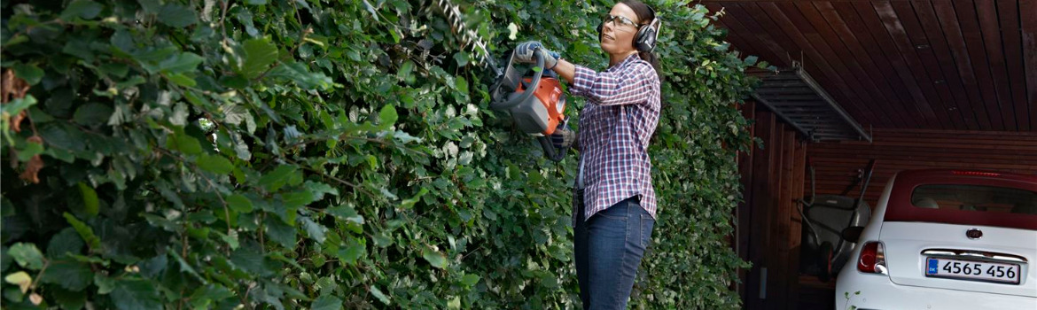 A woman wearing ear protection and work gloves uses a handheld hedge trimmer to cut leaves off of bushes next to her garage.